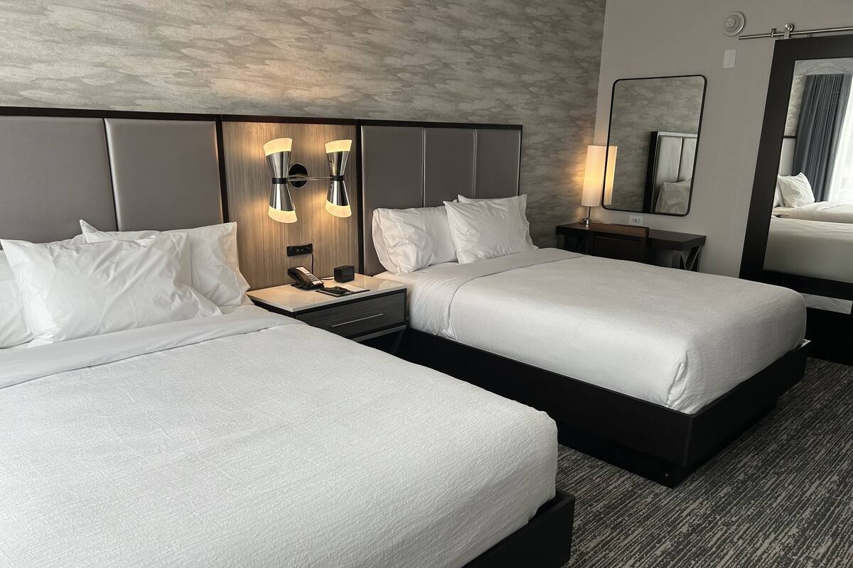 A hotel room with two beds, a nightstand with a phone and a lamp, a large mirror, and a textured wall.
