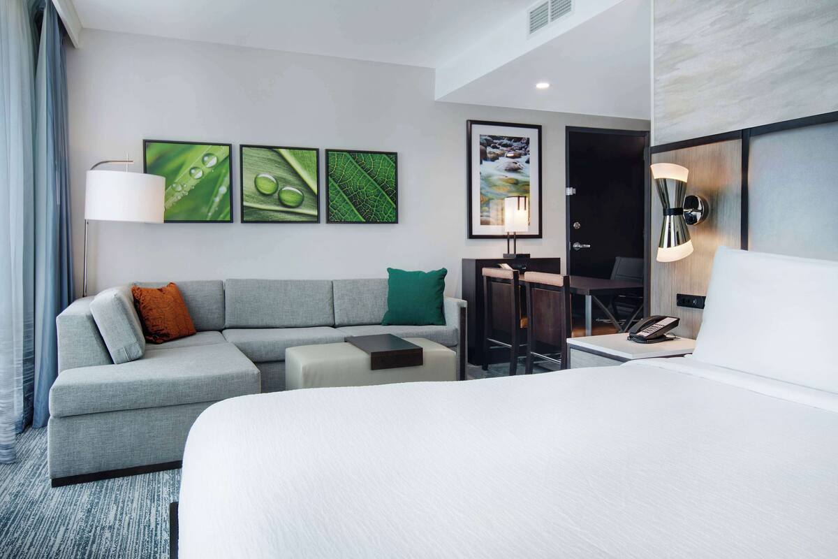 A modern hotel room with a bed, a grey sofa, a desk, and a chair. There are three framed pictures on the wall, a lamp, and a phone on the desk.