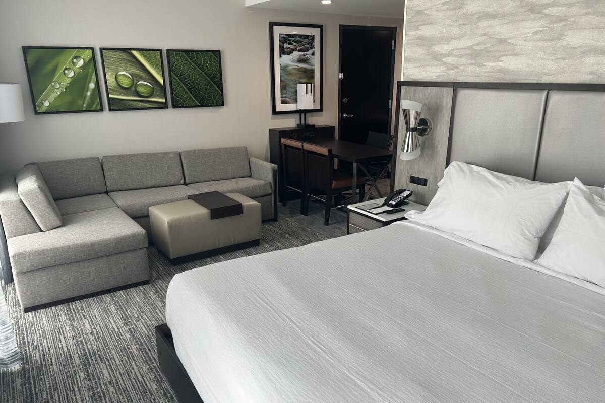 A modern hotel room with a large bed, a sofa, a desk, and a chair. There are three framed pictures on the wall.