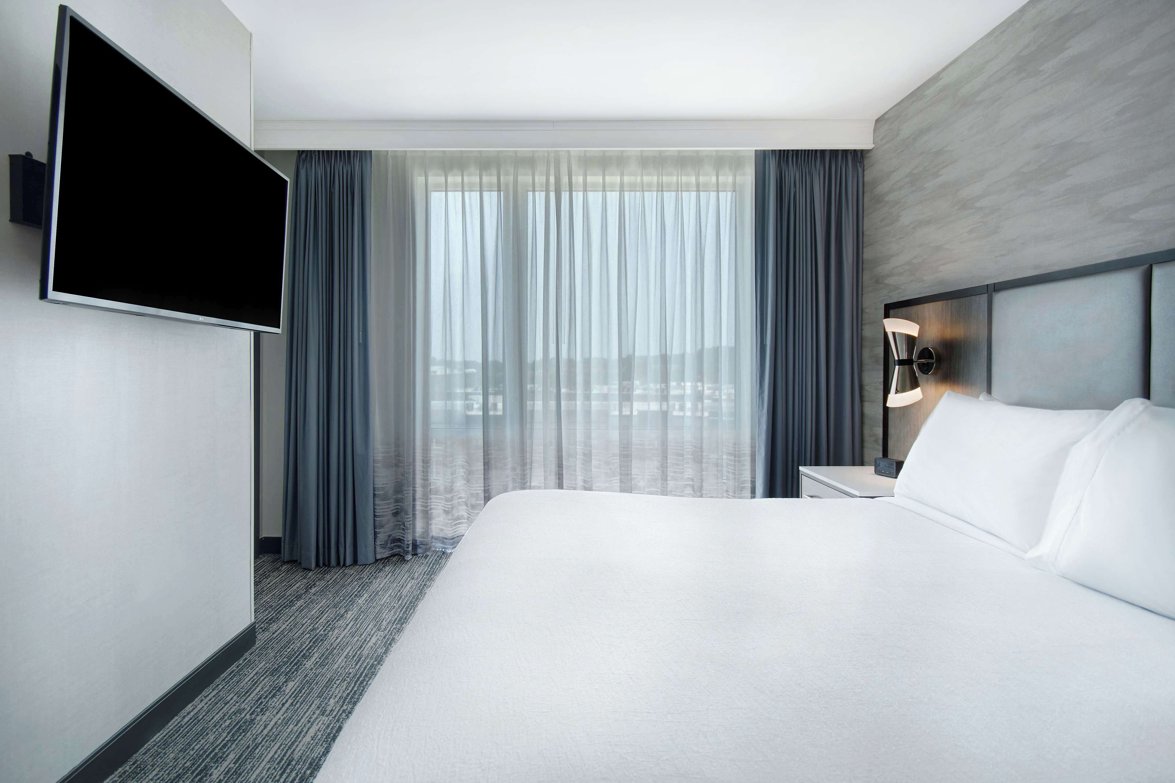 Een moderne hotelkamer met een groot bed, een flatscreen televisie aan de muur, een nachtkastje met een lamp, en uitzicht op de stad door dunne gordijnen.