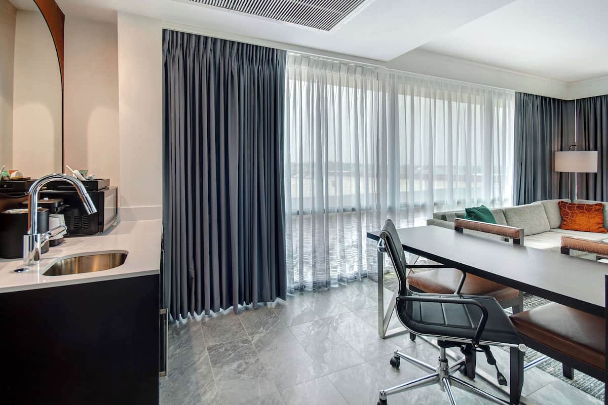 A modern hotel room with a dining area, a kitchen with a sink, and large windows with curtains.