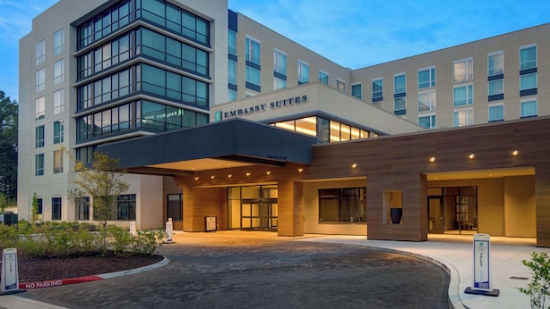 A modern building with a sign reading "Embassy Suites." It features a large entrance with glass doors, a covered area, and a landscaped area with plants. - Embassy Suites By Hilton Alpharetta Halcyon (Alpharetta)