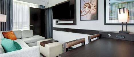 A modern hotel room with a grey sofa, a small table, a flat-screen TV mounted on the wall, and a painting above the TV.