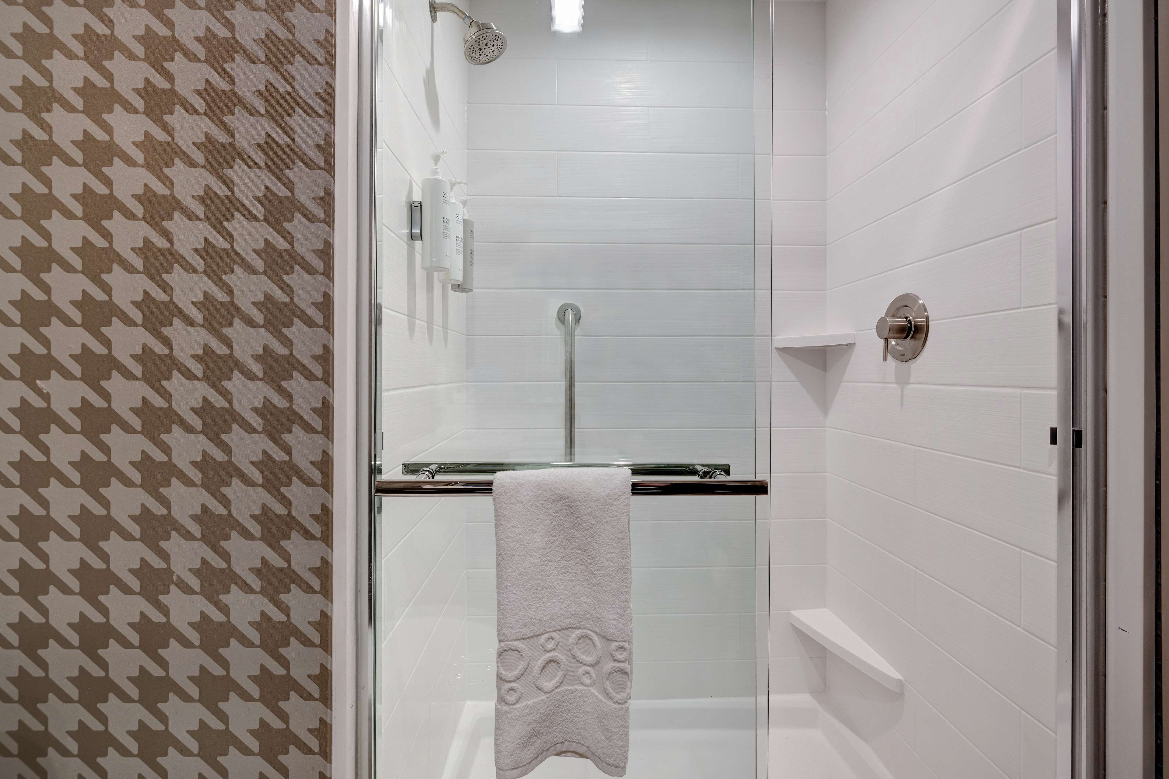 Zona de ducha con paredes de azulejos blancos, una puerta de cristal y una toalla colgada en un perchero.