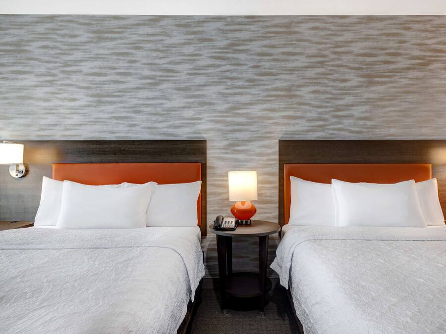 Quarto de hotel com duas camas, uma mesa redonda com abajur e parede texturizada.