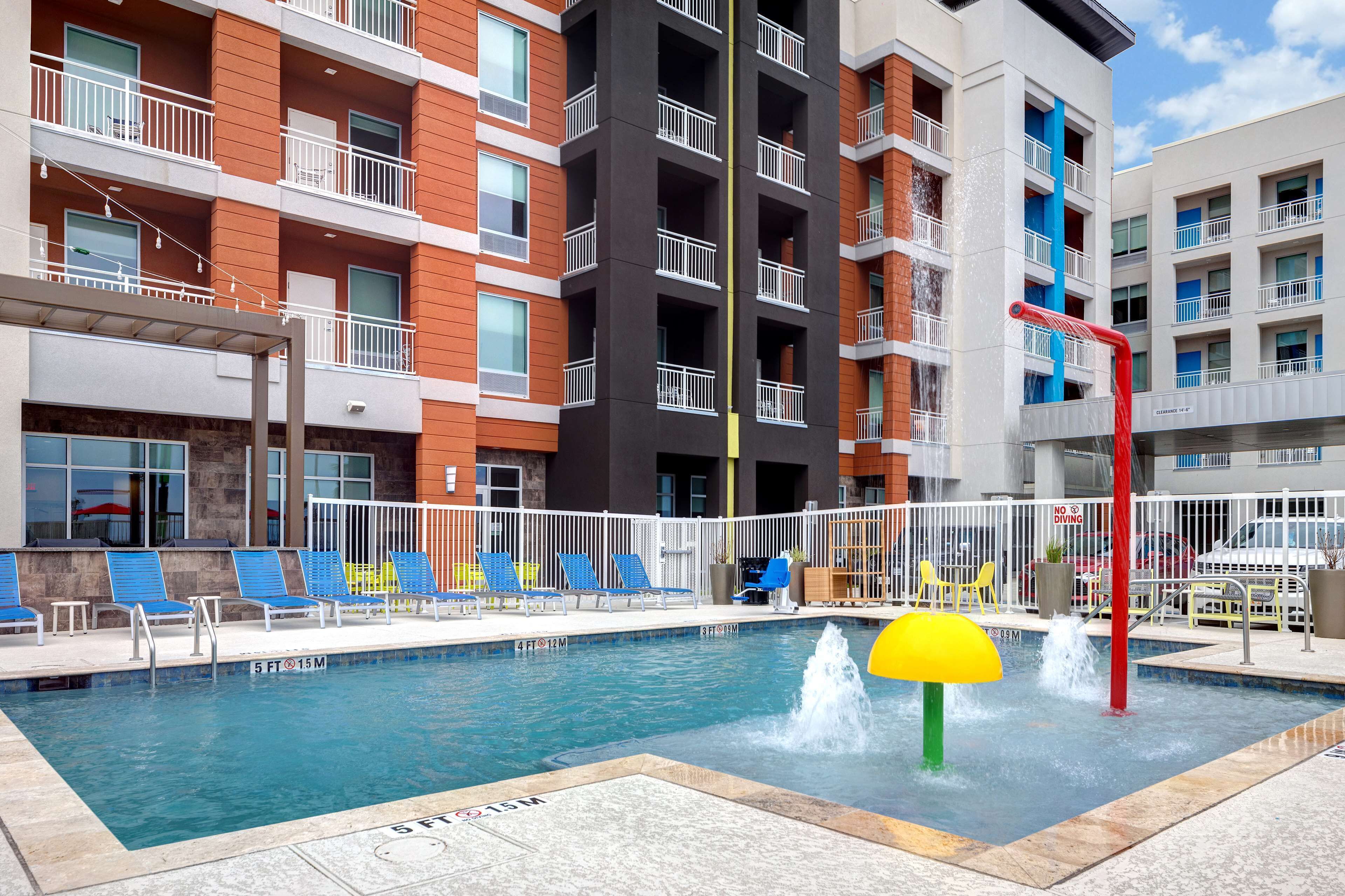 Une piscine avec des chaises longues bleues et jaunes, une fontaine et un panneau indiquant qu’il est interdit de plonger.