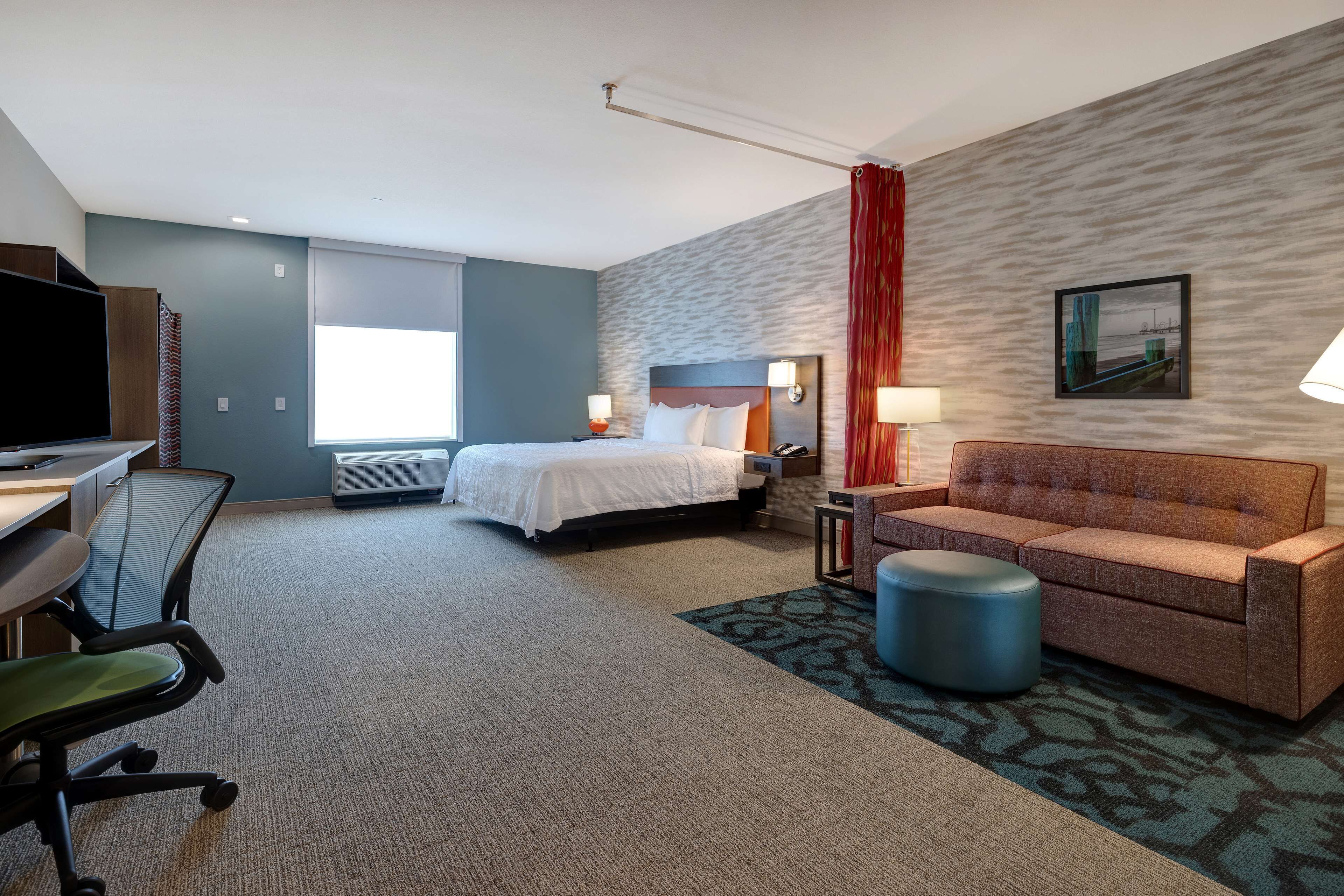 A hotel room with a bed, a desk with a chair, a sofa, a small table with a lamp, and a wall-mounted TV.