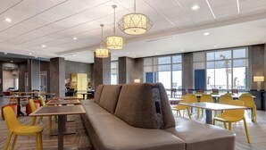 Een moderne eetruimte met een lange bank, gele stoelen en tafels. Er hangen hanglampen en in de achtergrond bevinden zich grote ramen.