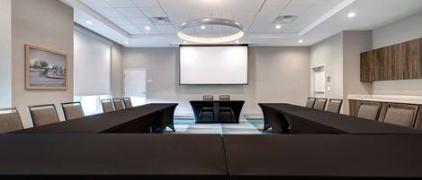A conference room with a long table, chairs, a projector screen, and a large painting on the wall.