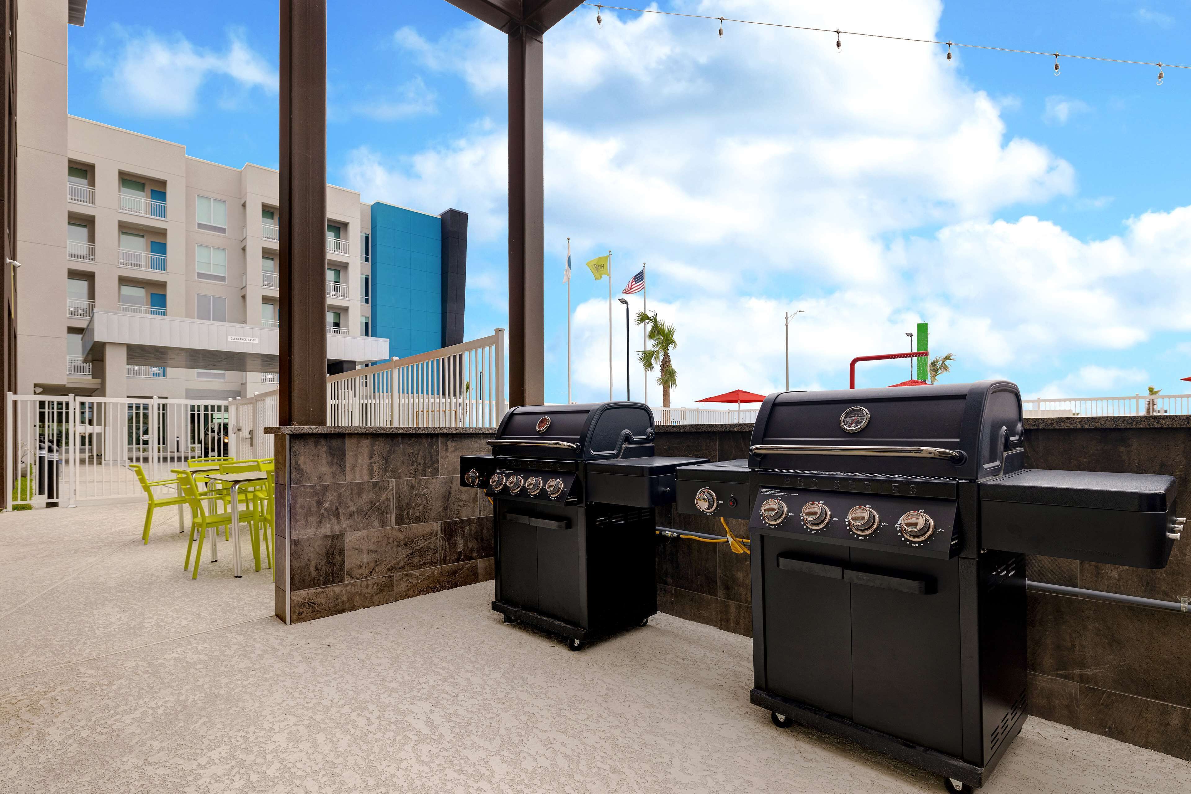 An outdoor patio with two black barbecue grills, a yellow table with chairs, and a building in the background.