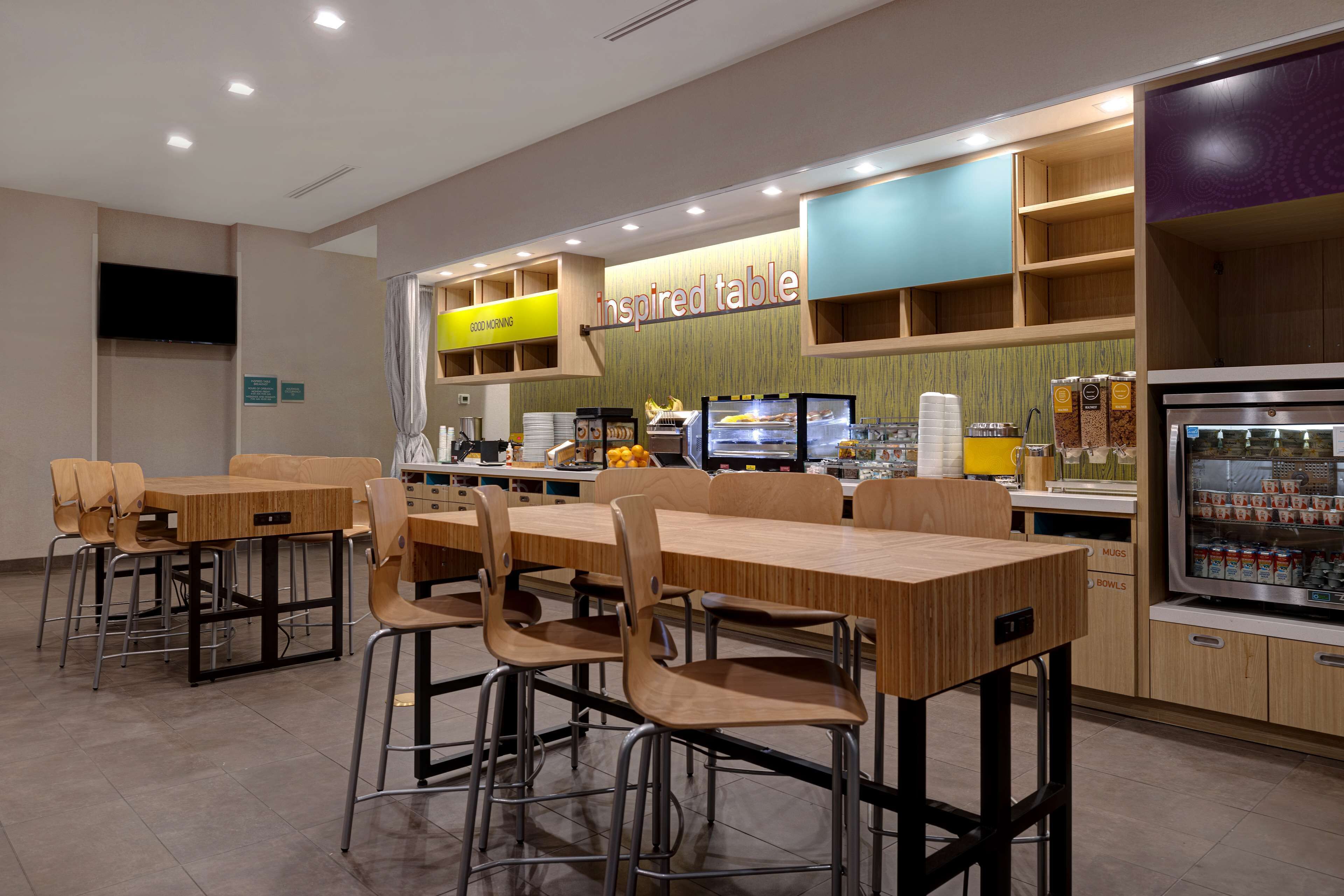 Een modern restaurant met houten tafels en stoelen, een toonbank met een menu, en een koelkast vol drankjes.