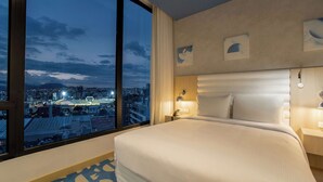 Hotellihuone, jossa on suuri sänky, yöpöytä, lamppu ja näkymä kaupungin siluettiin auringonlaskun aikaan.