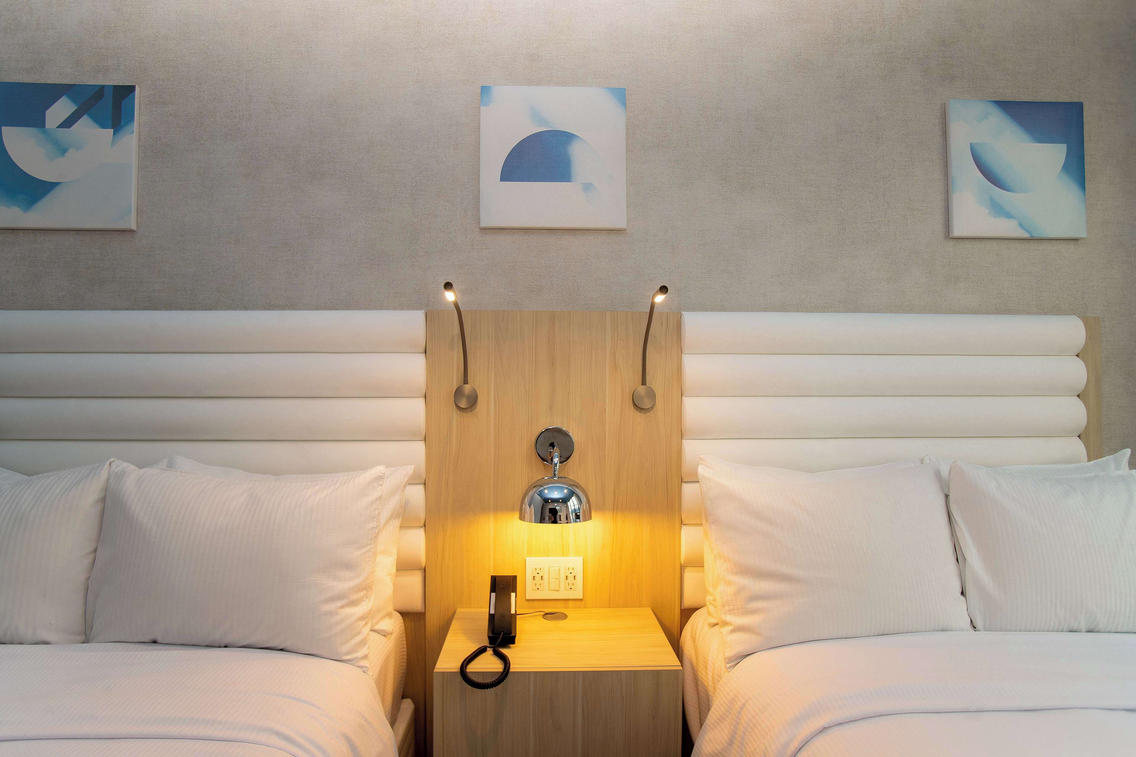 A hotel room with a wooden headboard, two beds with white bedding, a bedside table with a lamp, and abstract art on the walls.