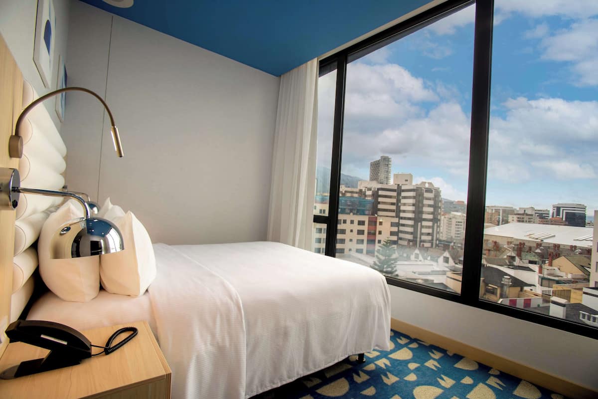A hotel room with a bed, a desk with a lamp, a telephone, and a large window offering a city view.