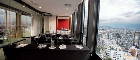 Un salón de conferencias con una mesa negra preparada para una reunión, ventanas amplias que ofrecen vistas a la ciudad y una pared de acento roja.