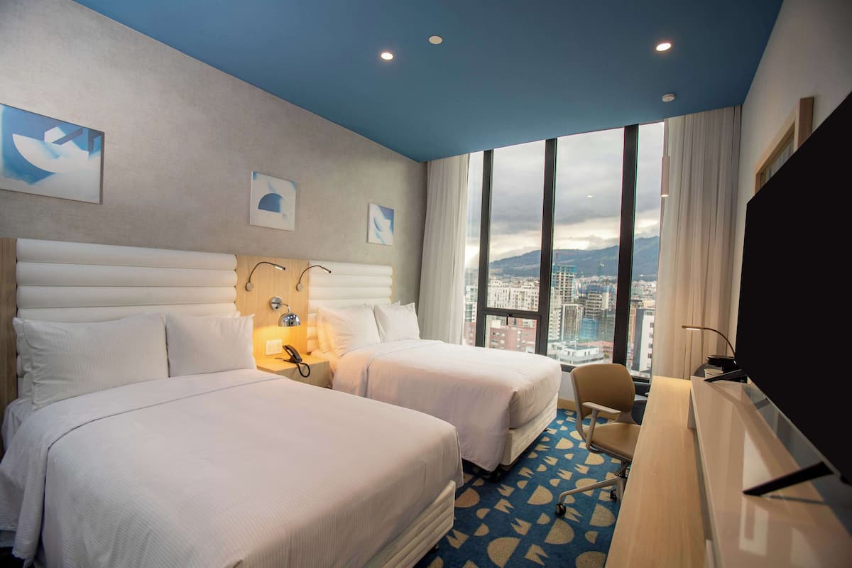 A hotel room with two beds, a desk, a chair, and a large window offering a city view.