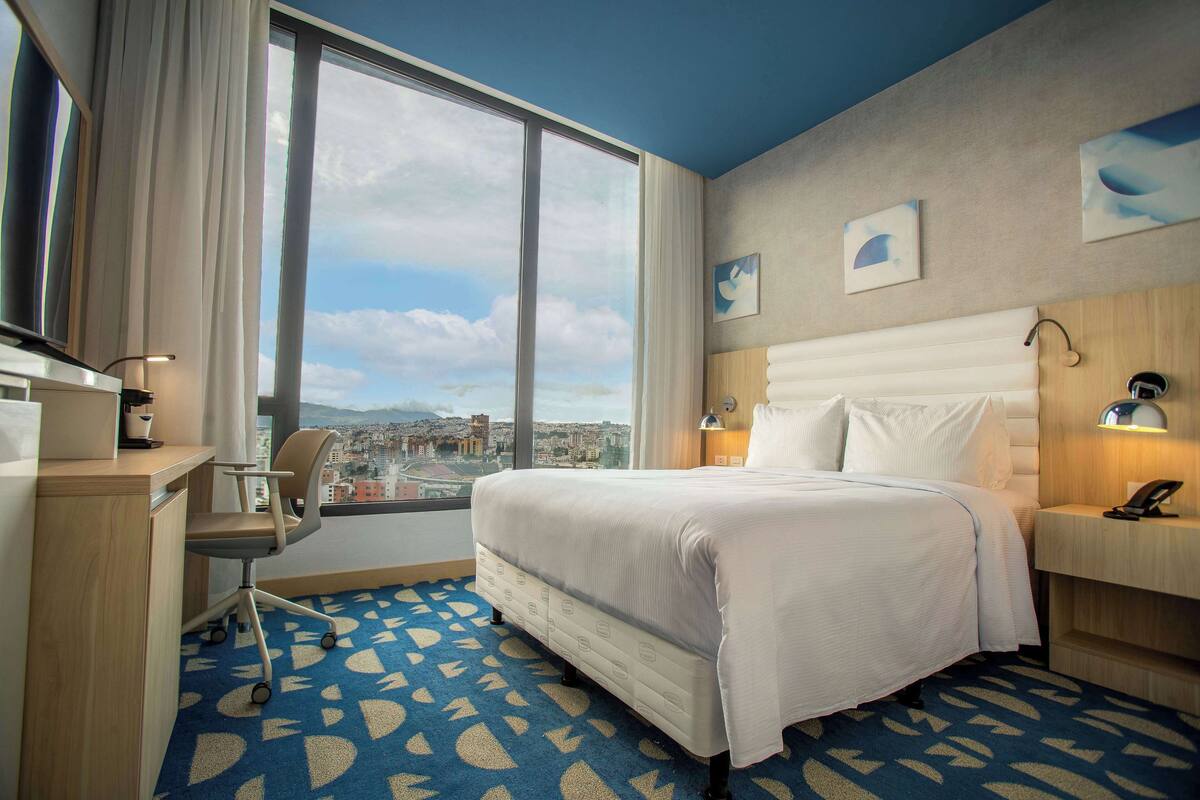 A hotel room with a large window, a bed, a desk, and a chair.