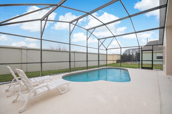 Una piscina con cerramiento de vidrio, rodeada por una terraza de hormigón y sillas reclinables blancas.
