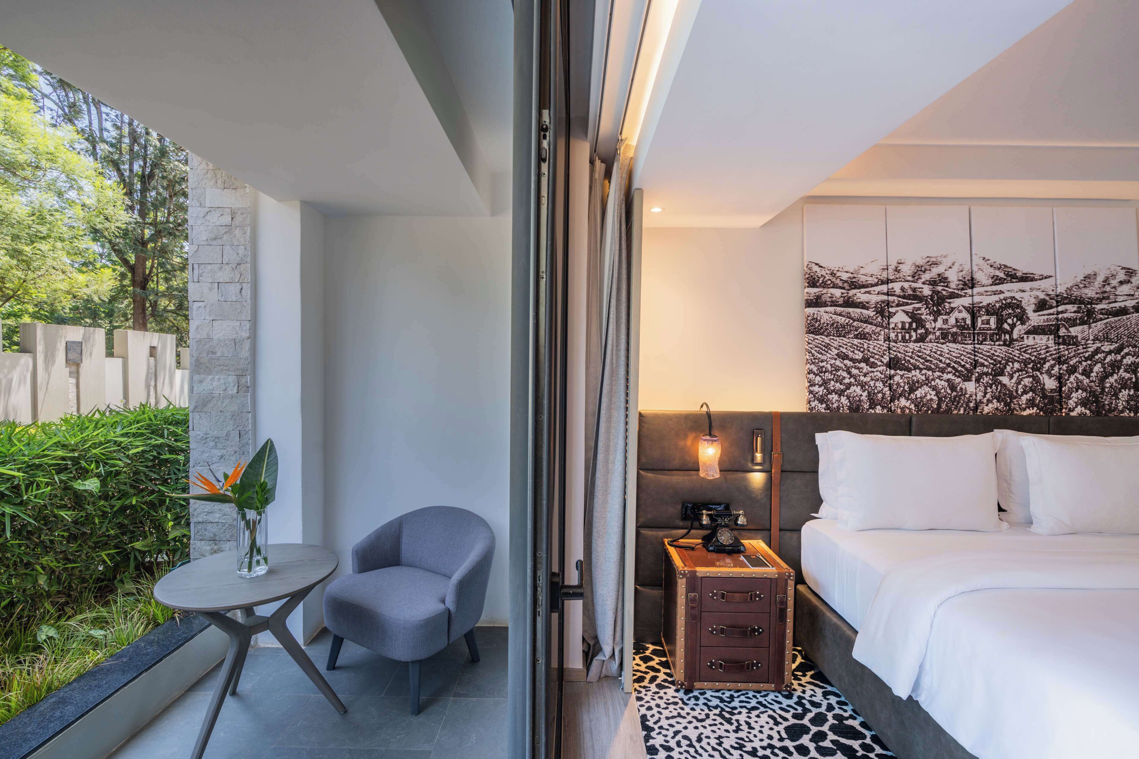 Une chambre d’hôtel moderne, équipée d’un lit, d’une petite table avec une chaise, d’une table de chevet avec une lampe, et offrant une vue sur la verdure environnante.