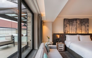 A modern hotel room with a large bed, a sofa, a small table with a lamp, and a view of the outside balcony.