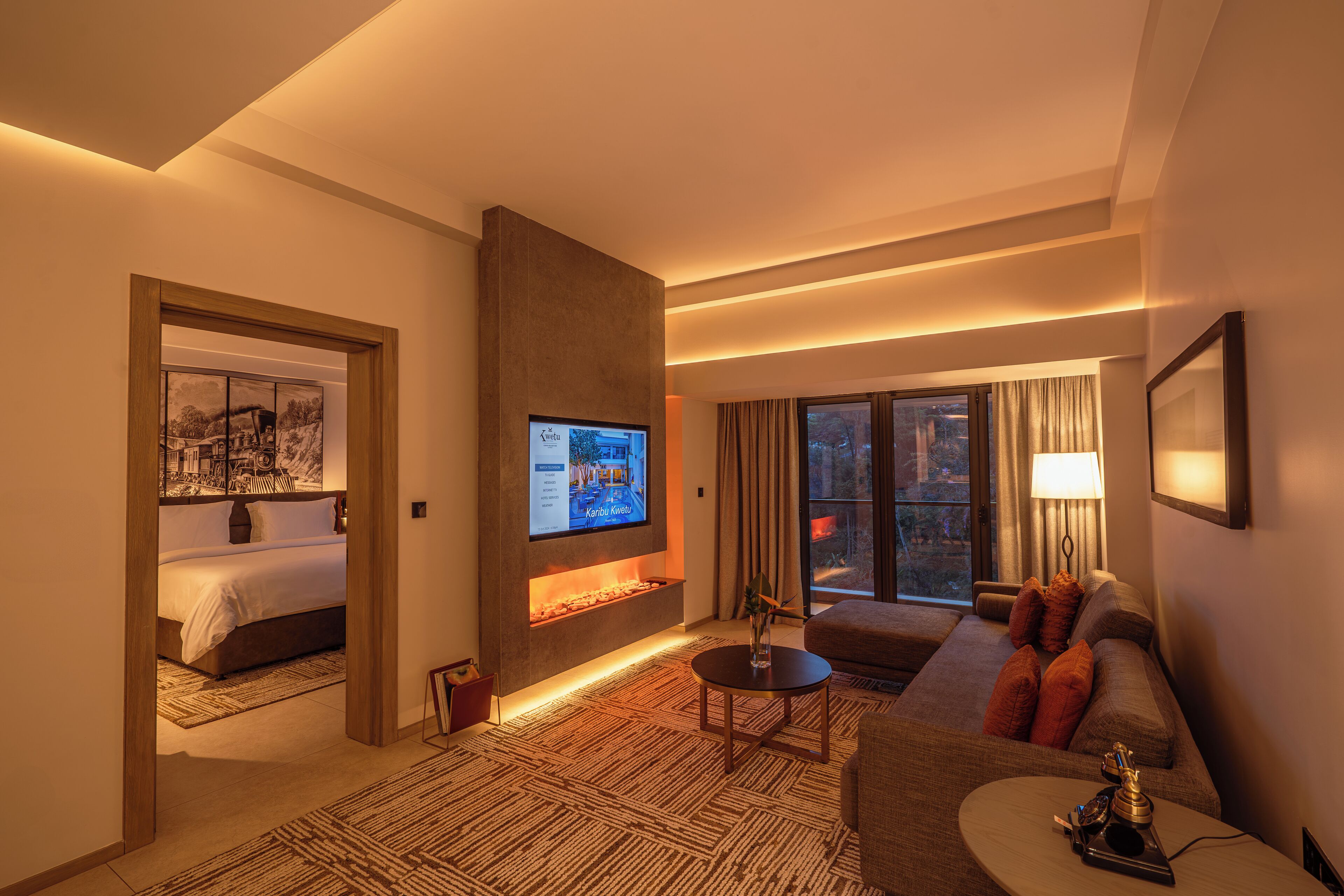 A modern hotel room with a large bed, a flat-screen TV, a fireplace, a sofa, and a dining area.