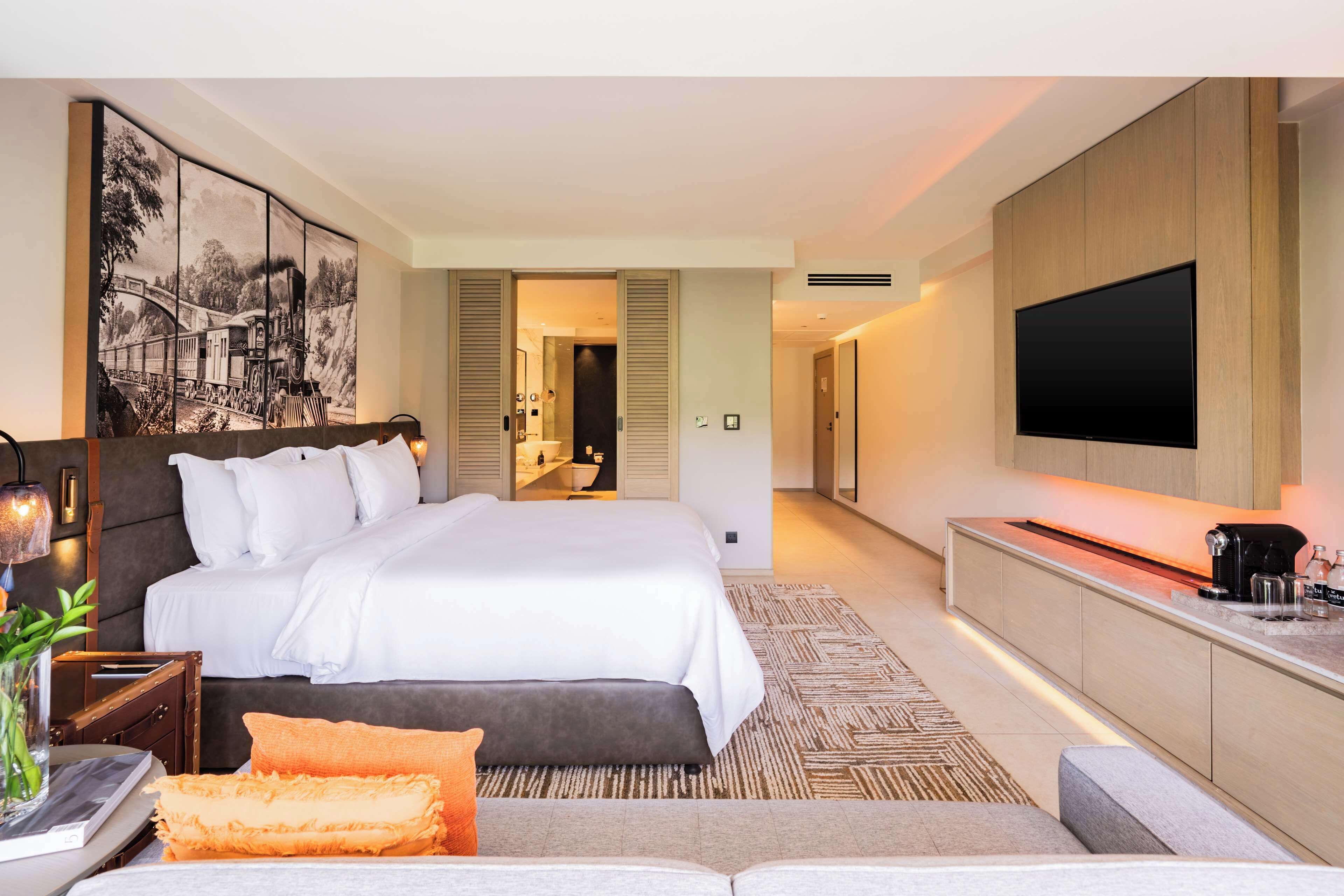 A modern hotel room with a large bed, a flat-screen TV, a bedside table with a lamp, and a painting on the wall.
