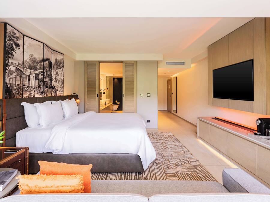 Una habitación de hotel moderna con una cama grande, un televisor de pantalla plana, una mesita de noche con lámpara y un cuadro en la pared.