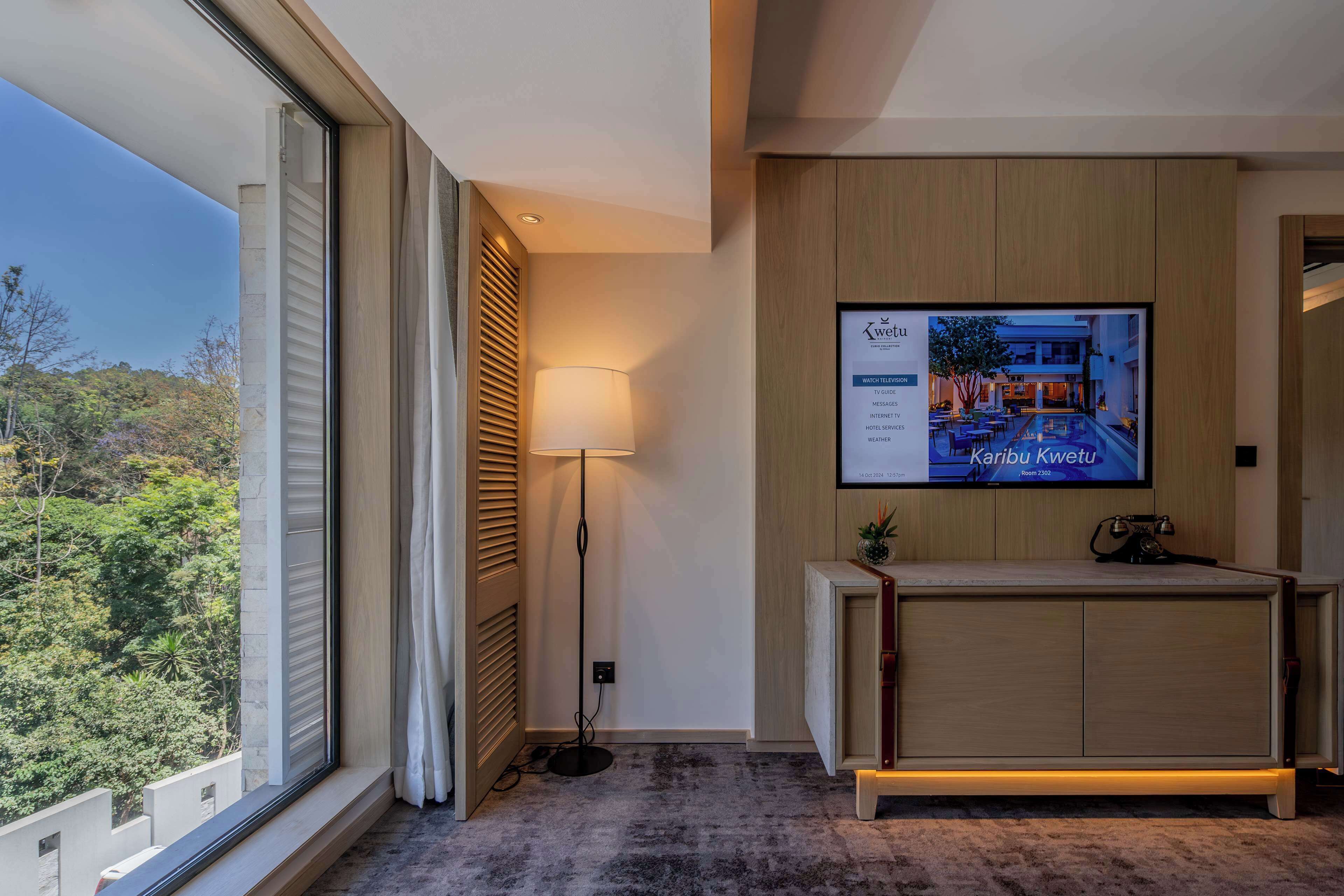 A modern hotel room with a large window, a floor lamp, a reception desk, and a TV displaying the hotel's name and amenities.