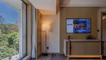 A modern hotel room with a large window, a floor lamp, a reception desk, and a TV displaying the hotel's name and amenities.