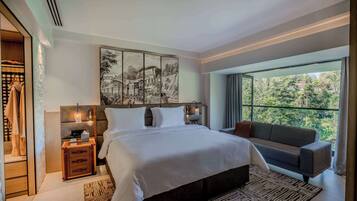 Une chambre moderne avec un grand lit, un canapé, une table de chevet avec une lampe, et une vue sur la verdure grâce à de grandes fenêtres.