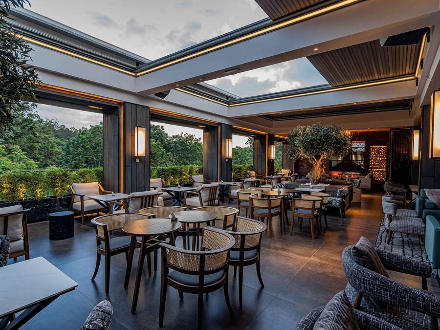 An indoor-outdoor dining area with multiple tables and chairs, large windows, and a view of greenery.
