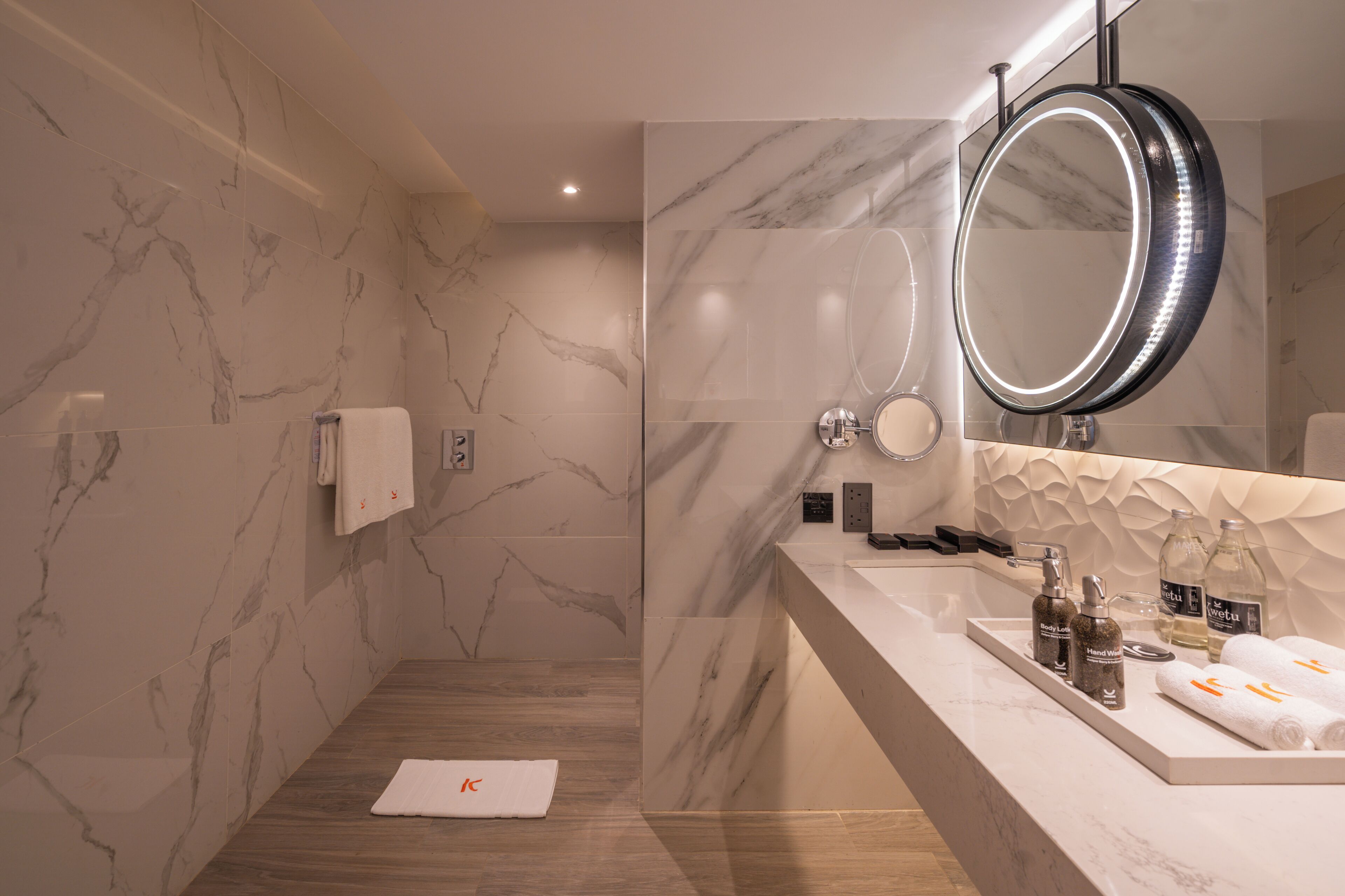 A modern bathroom with marble walls, a wooden floor, a large mirror with LED lights, and a vanity with toiletries.