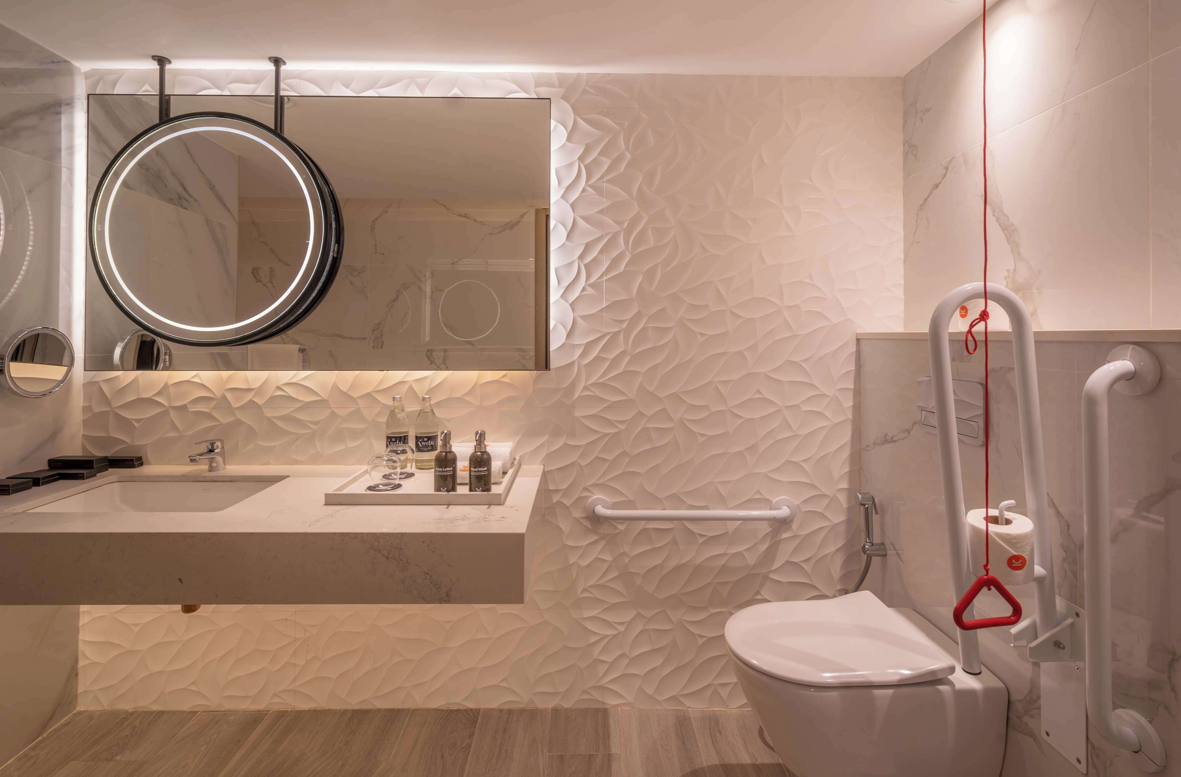 A modern bathroom with a marble vanity, a round mirror, and a toilet with a safety grab bar.