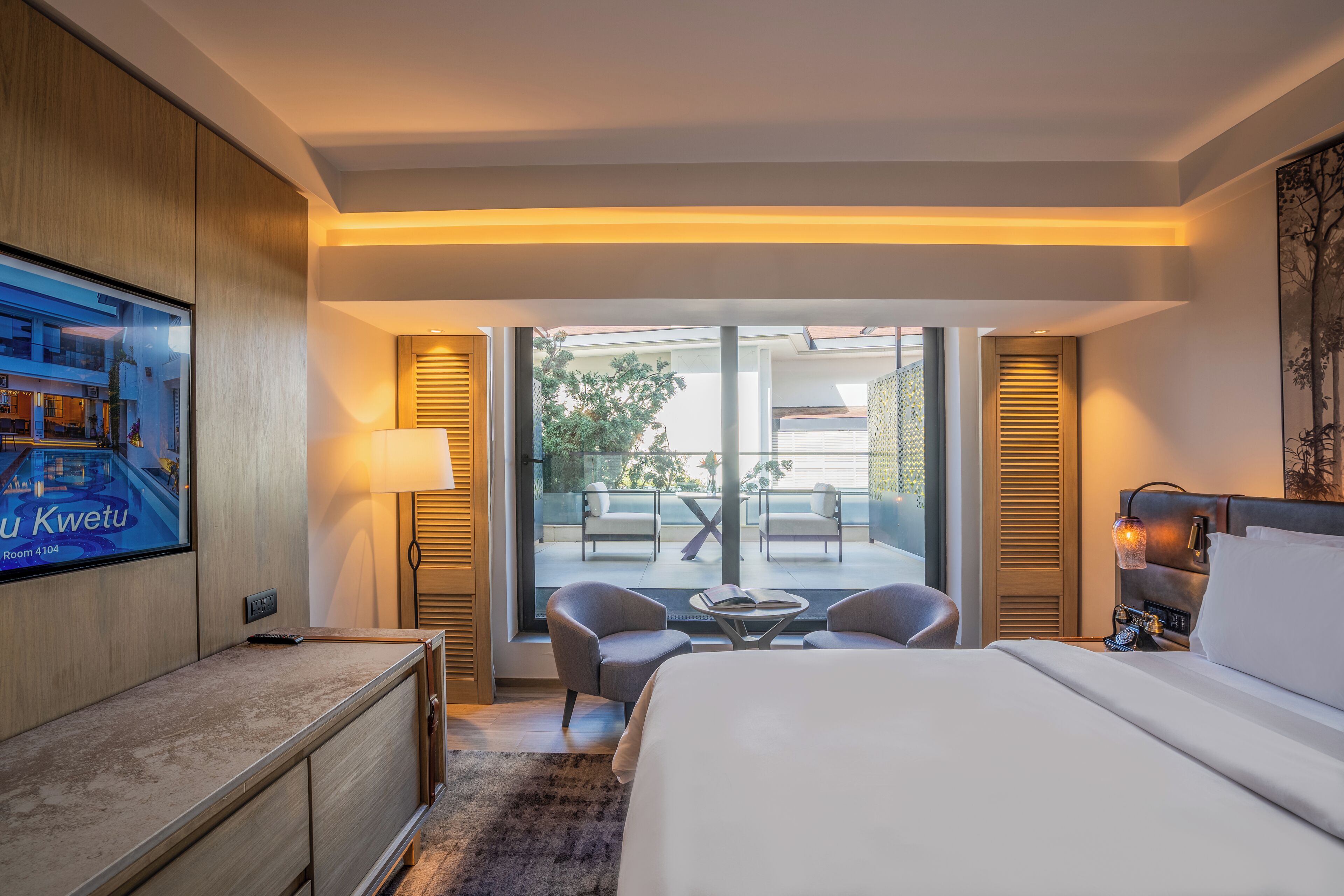 Habitación de hotel con cama, escritorio, televisión, balcón con sillas y vistas al exterior.