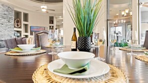 Un coin repas avec une table ronde dressée pour deux personnes, agrémentée de sets de table en fibres tressées, d’une bouteille de vin et d’un vase contenant des feuilles vertes.