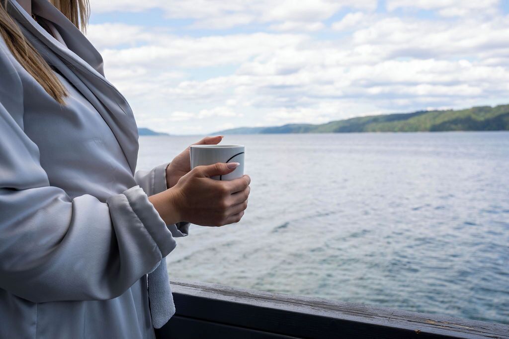 A person holding a mug, overlooking a body of water with a distant landmass.