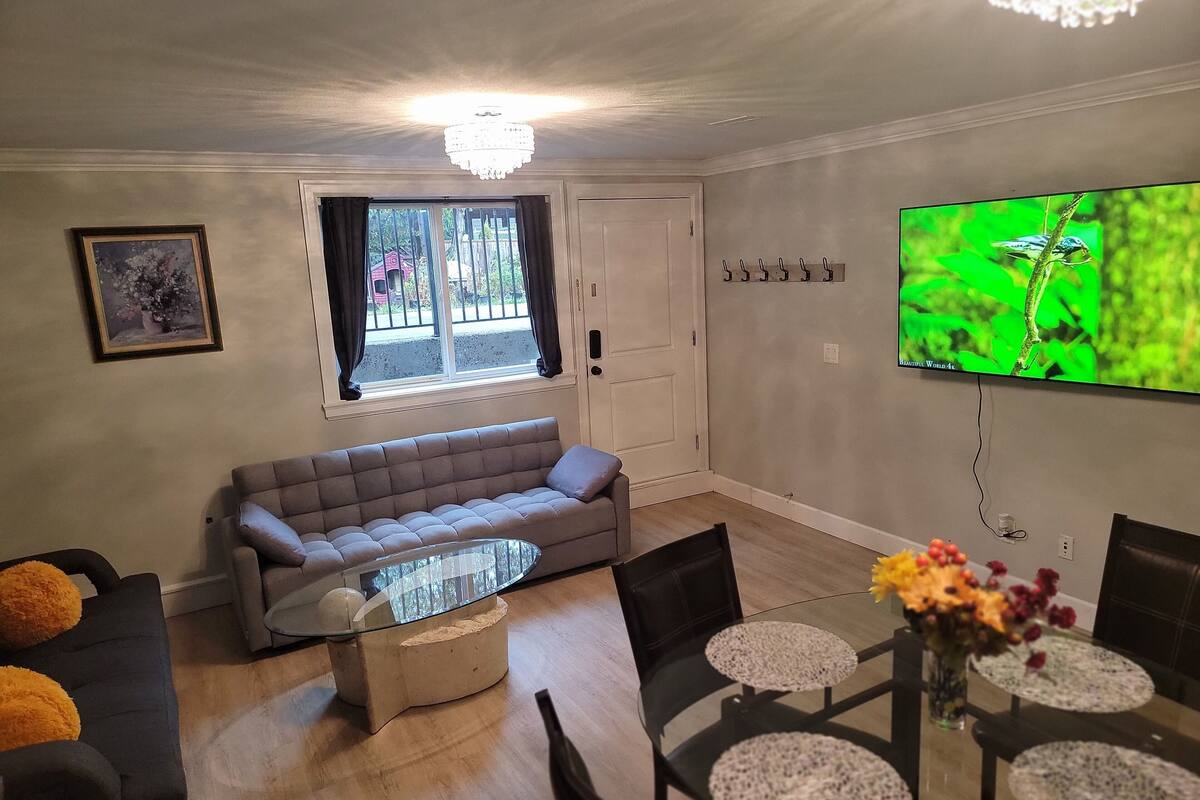 A living room with a sofa, a glass coffee table, a dining table with chairs, a TV mounted on the wall, and a window with curtains.