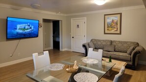 A living room with a glass dining table, a sofa, a television mounted on the wall, and a door leading to another room.