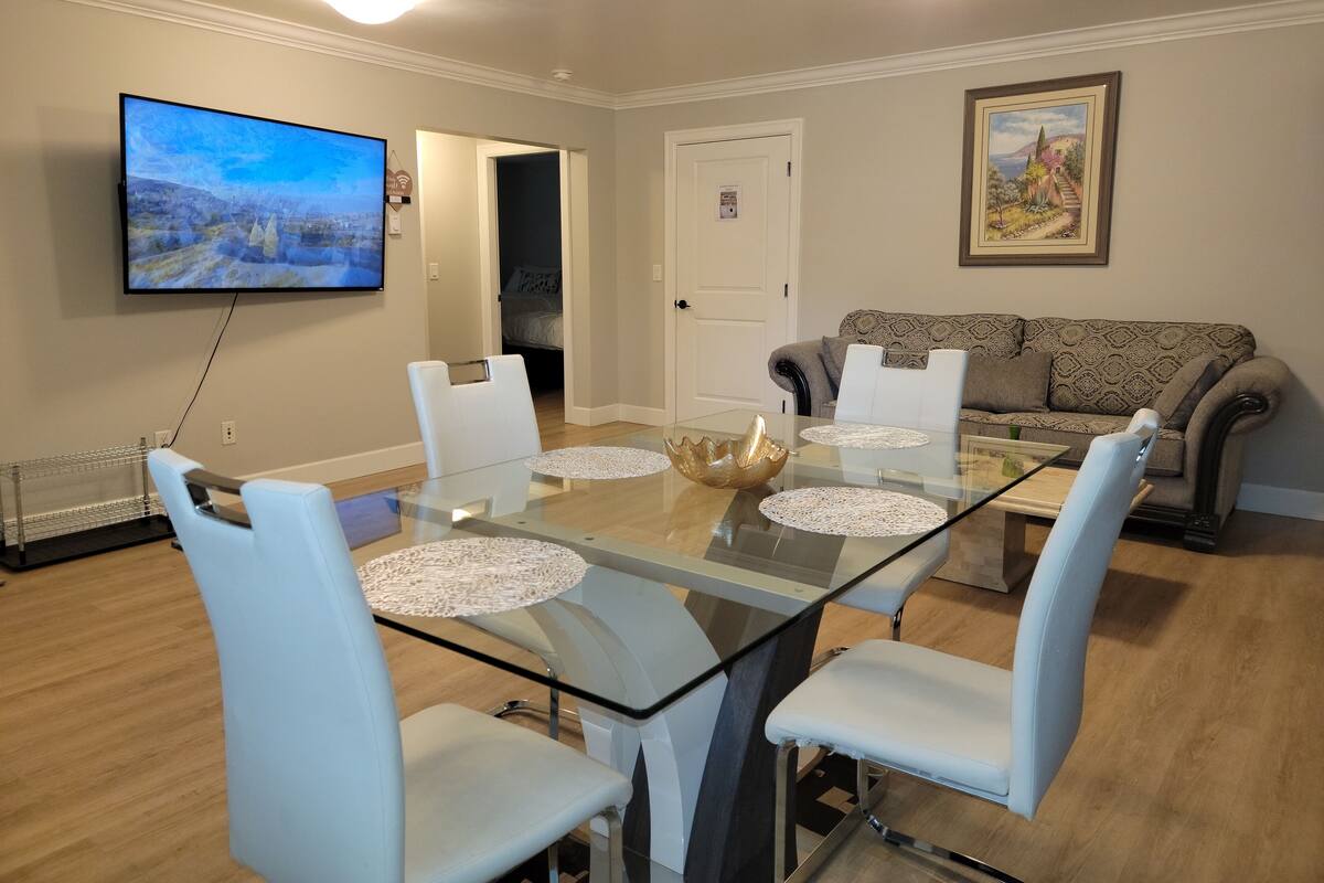 A dining area with a glass table, white chairs, a television, a sofa, and a door.