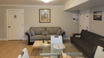 A living room with a patterned sofa, a glass coffee table, and a dark sofa. There are two doors, one with a sign and another with a picture.