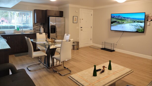 A modern kitchen with a stainless steel refrigerator, a dining area with a glass table and white chairs, and a flat-screen TV mounted on the wall.