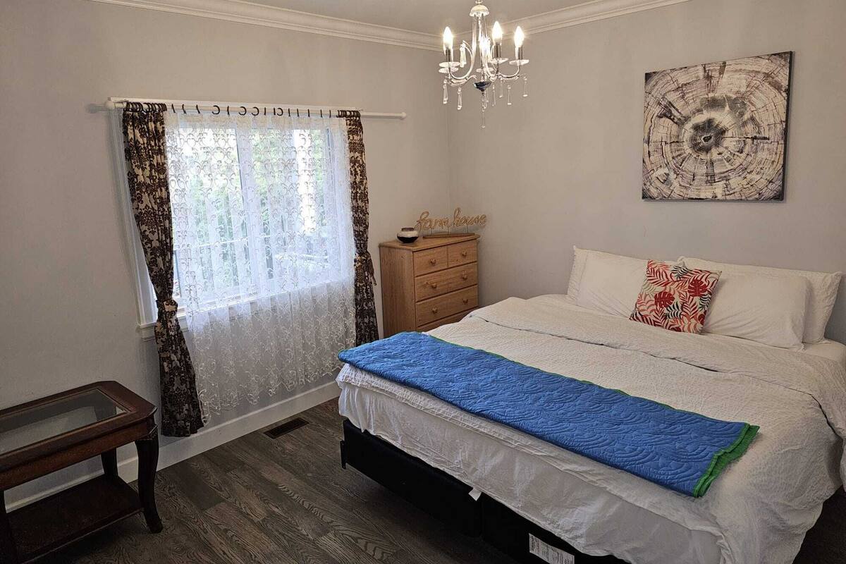 A bedroom with a bed, a chandelier, a wooden dresser, a bench, and a window with curtains.