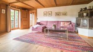 Een gezellige woonkamer met een houten plafond, een roze bank, een glazen salontafel en een kleurrijk vloerkleed.