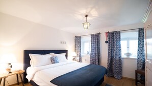 Un dormitorio ordenado con una cama grande, mesitas de noche, una lámpara colgante, cortinas con estampado y vistas al exterior.