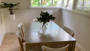 A dining area with a wooden table and white chairs, a potted plant, and a window with a view of greenery. - Amazing location across the road from Mount Martha South Beach Check in 10am (Mount Martha)