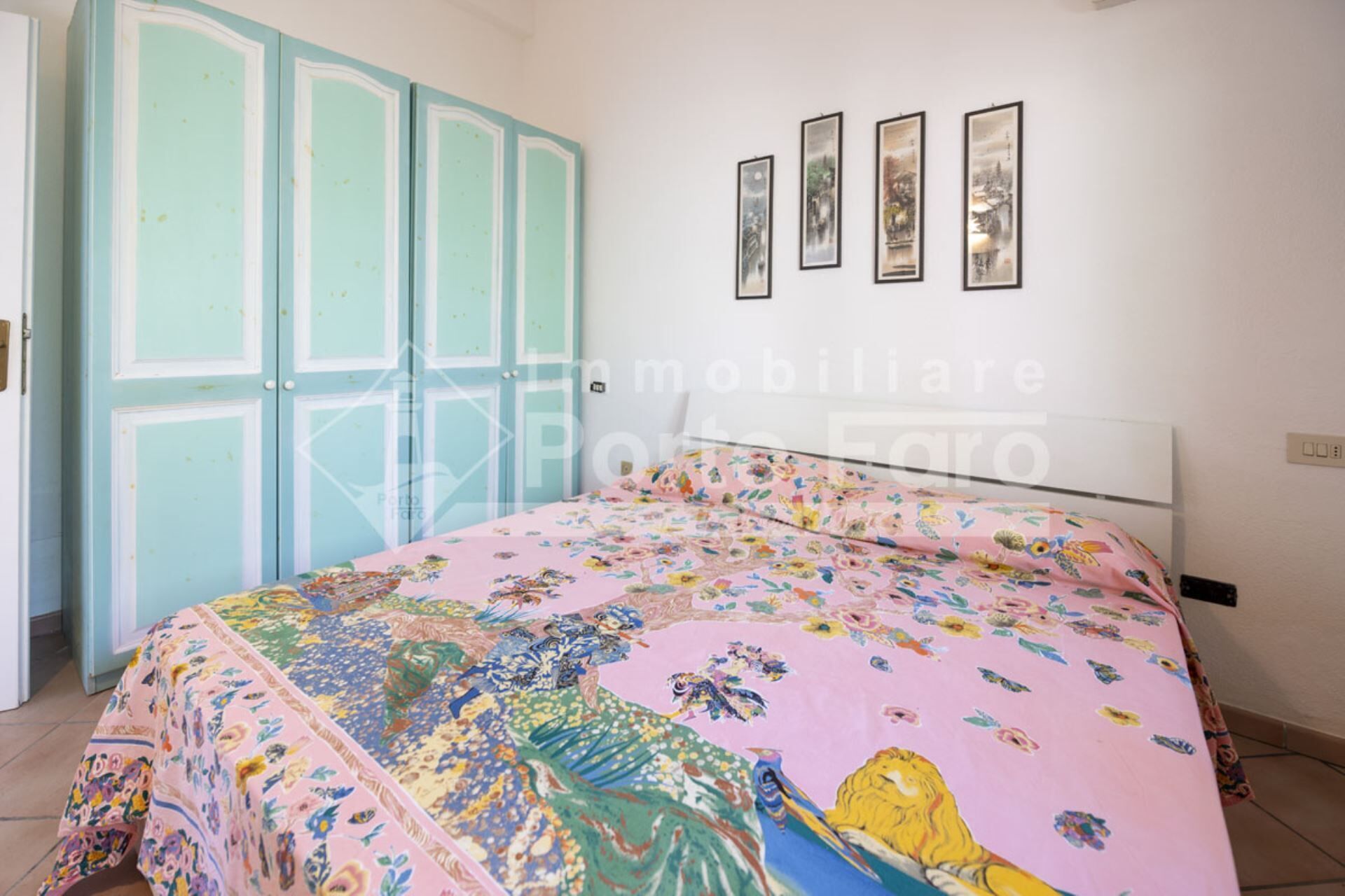 A bedroom with a bed covered in a floral-patterned bedspread, a teal-colored wardrobe, and four framed artworks on the wall.