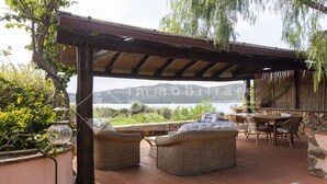 Un patio exterior con mobiliario de mimbre, una pérgola y vistas a un lago y montañas.