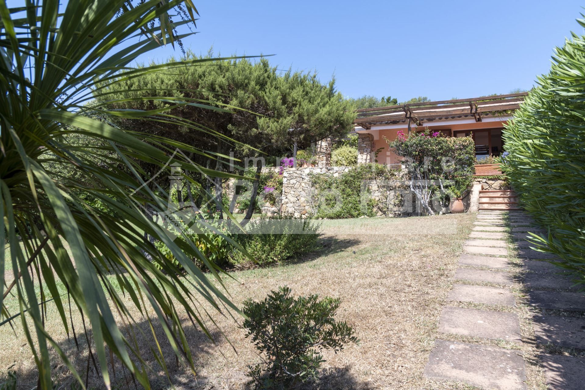 Un jardin avec un mur en pierre, un petit arbuste et un bâtiment doté d’un toit en tuiles.