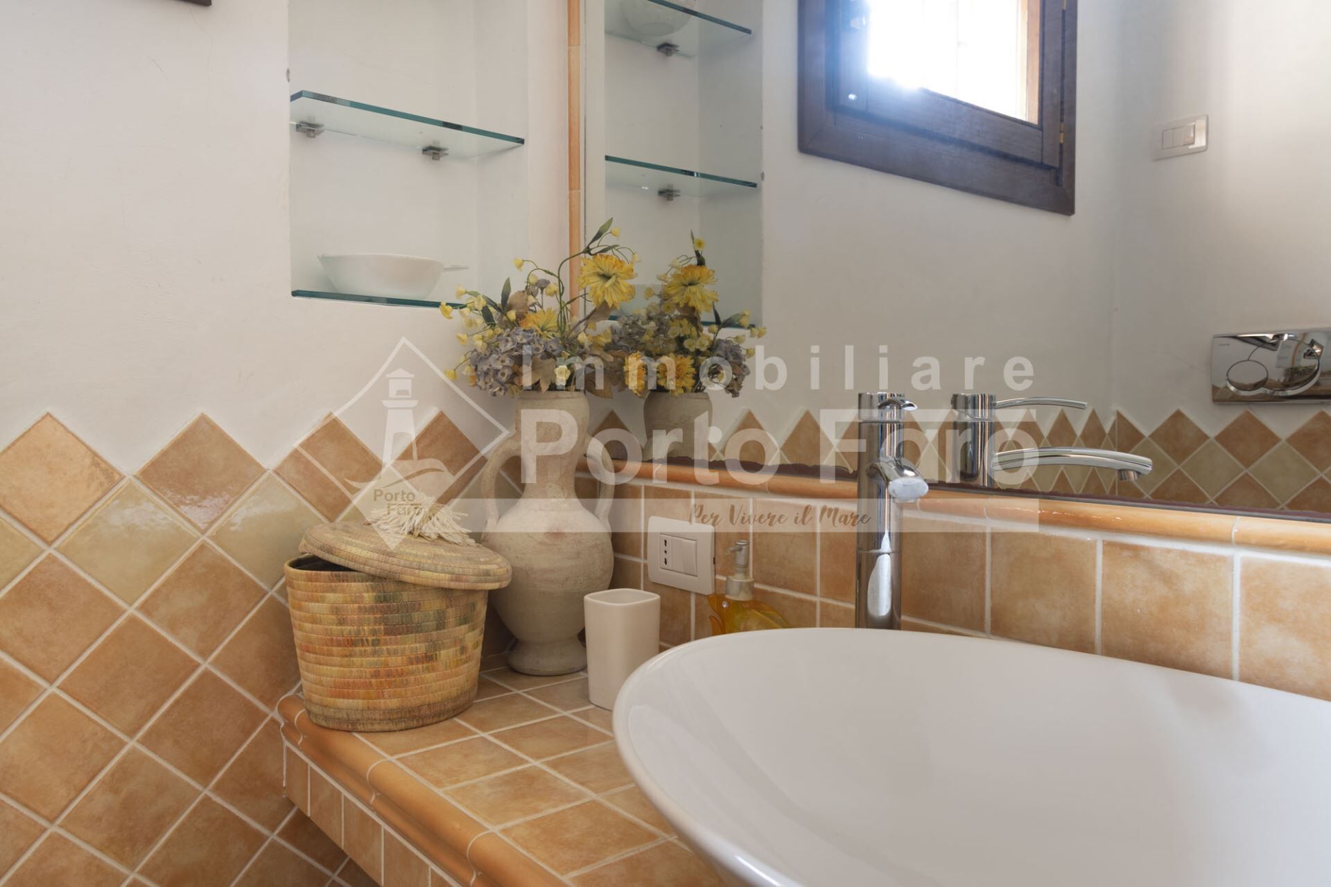 Un baño con un lavamanos blanco, un espejo y un florero con flores. Hay estantes encima del espejo y un perchero a la izquierda.