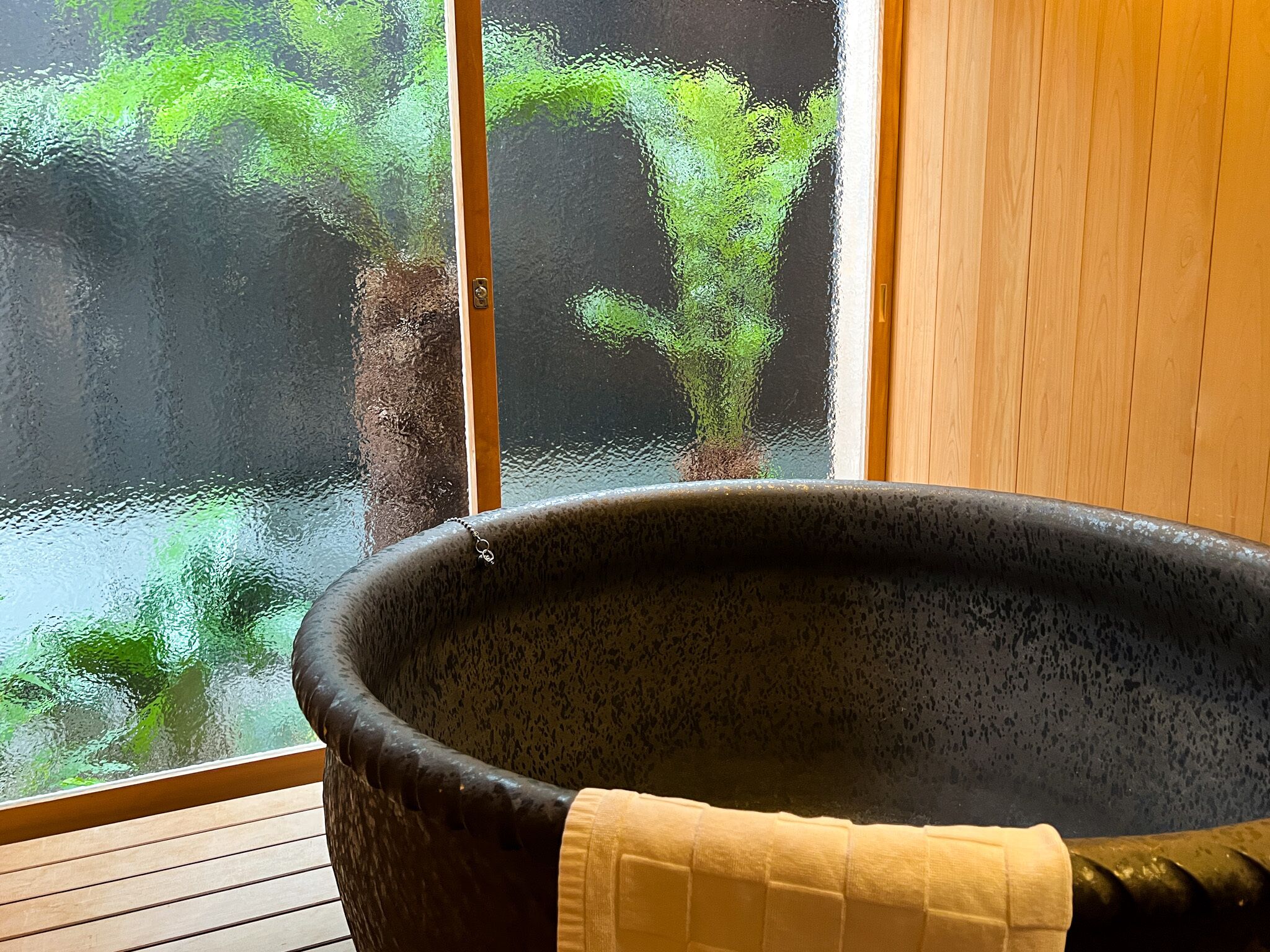 Un bain à remous en pierre, avec une serviette posée sur le bord, placé à côté d’un plancher en bois et d’une fenêtre donnant sur un paysage verdoyant.